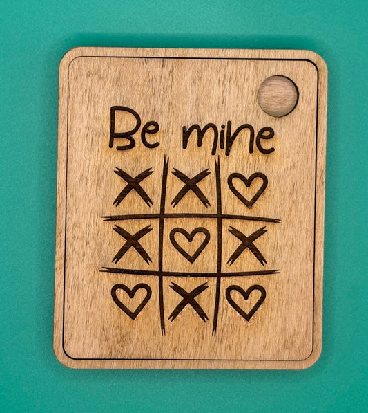 Wooden tic-tac-toe board with 'Be mine' text and heart symbols on a teal background
