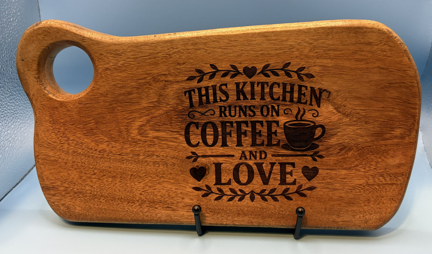 Wooden cutting board with engraved text 'This kitchen runs on coffee and love' on a light blue background.