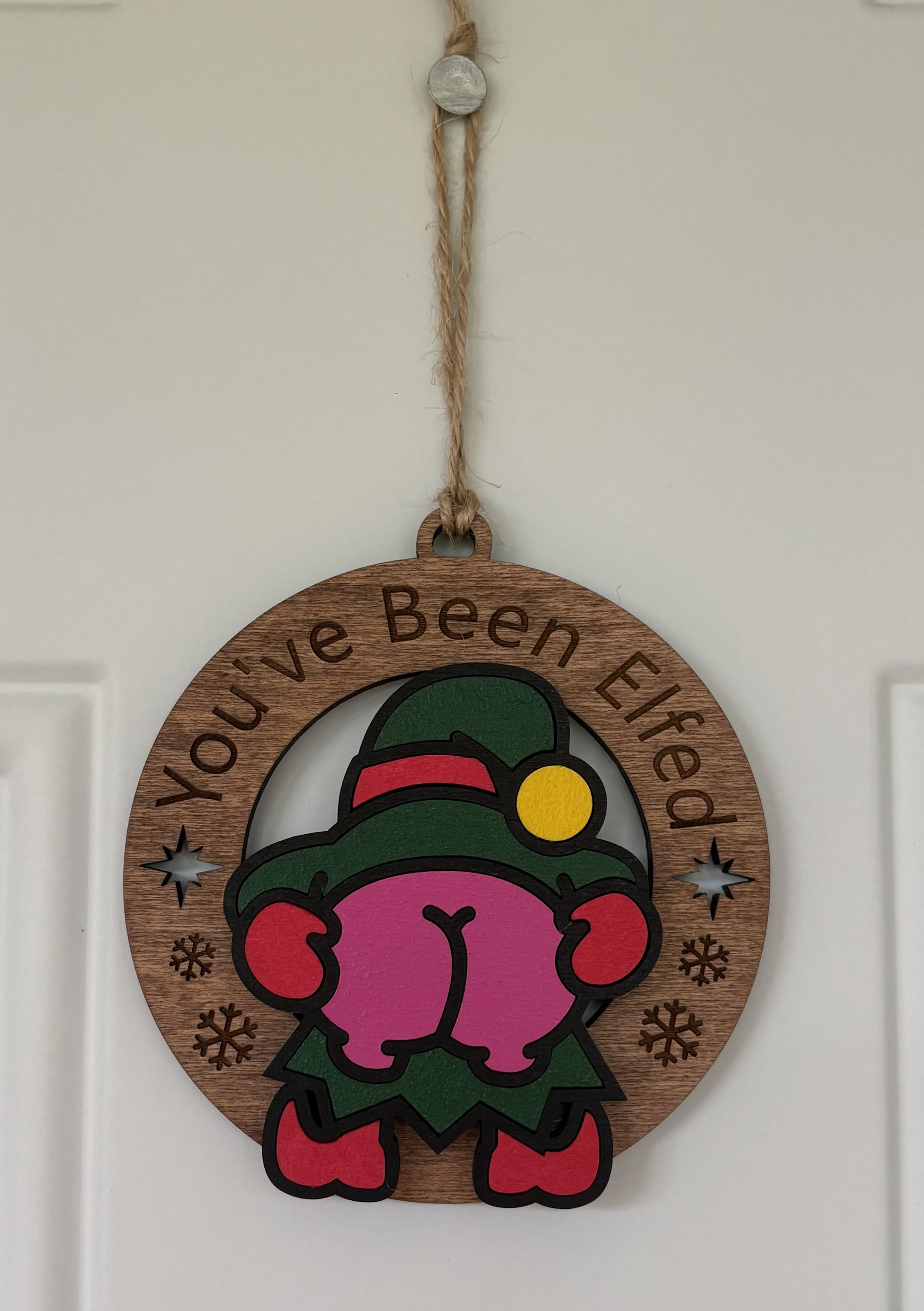 A round wooden hanging ornament with a jute rope, featuring a cartoon elf with a pink butt, green hat, and red shoes, with the text 'You've Been Elfed' on a white door.