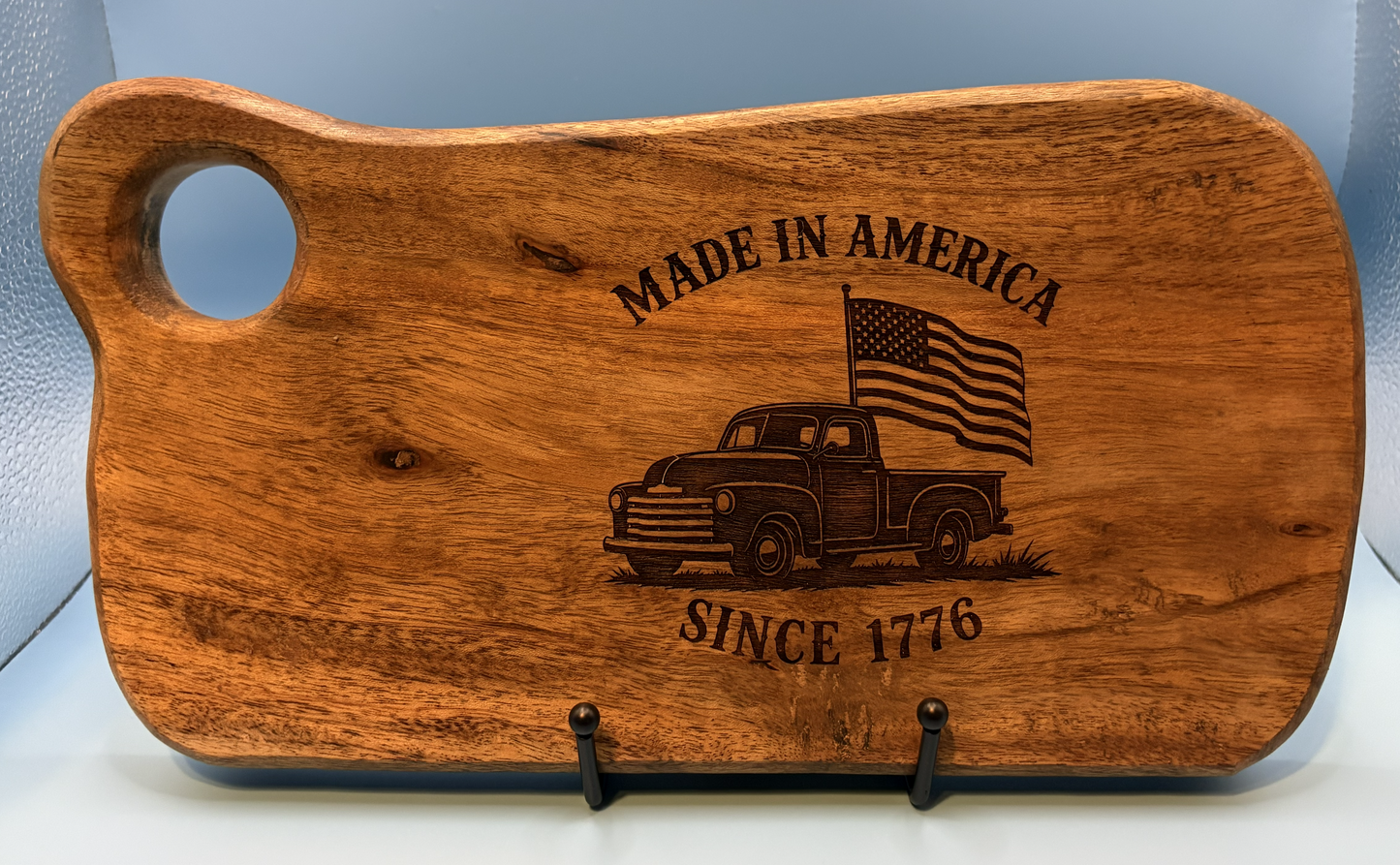 Wooden cutting board with engraved design of a truck and American flag, text 'Made in America Since 1776' on a light blue background.