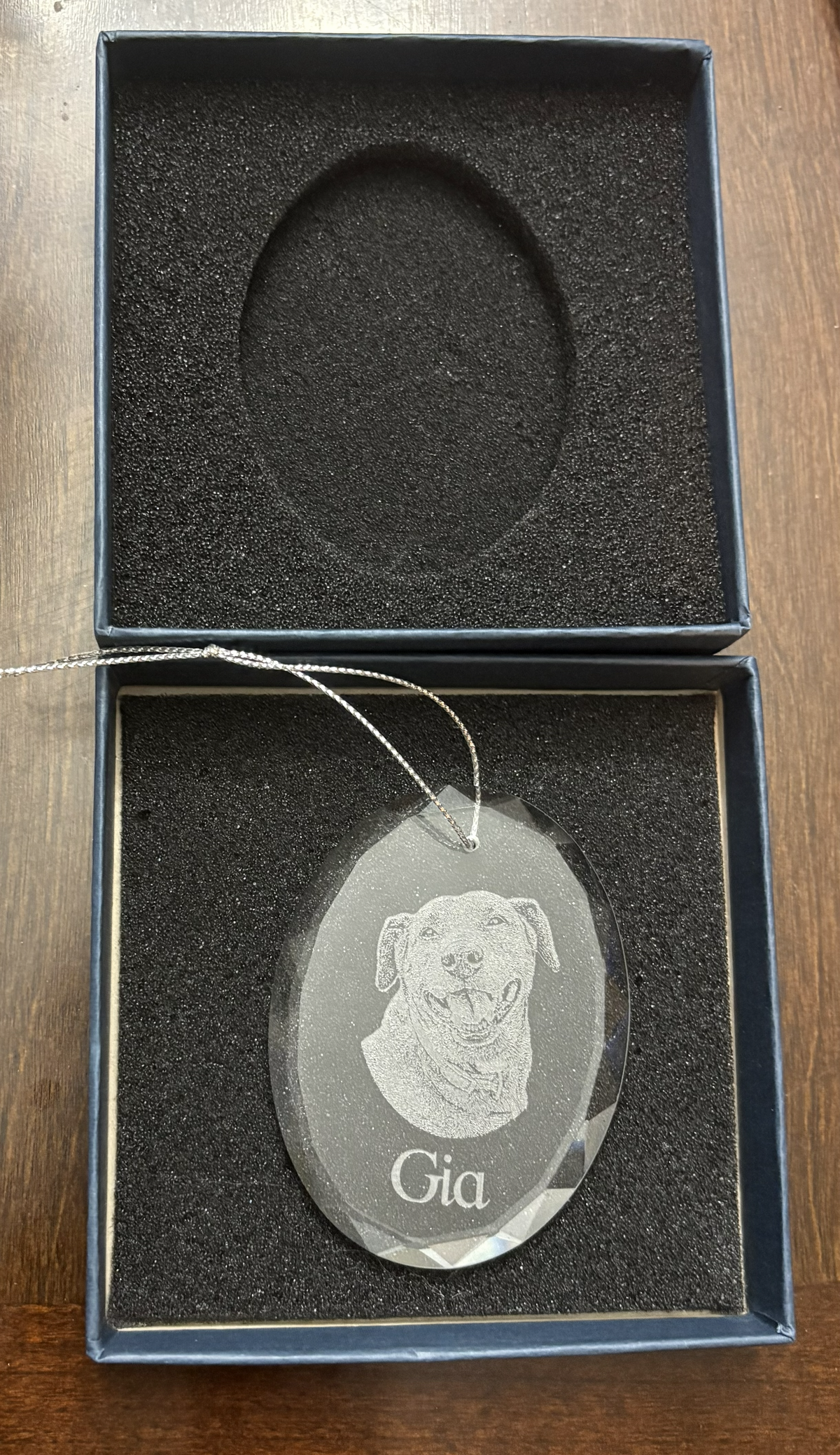 An oval personalized glass pet memorial ornament engraved with a dog portrait and the name 'Gia', presented in an open dark blue gift box with black foam insert on a wooden table.