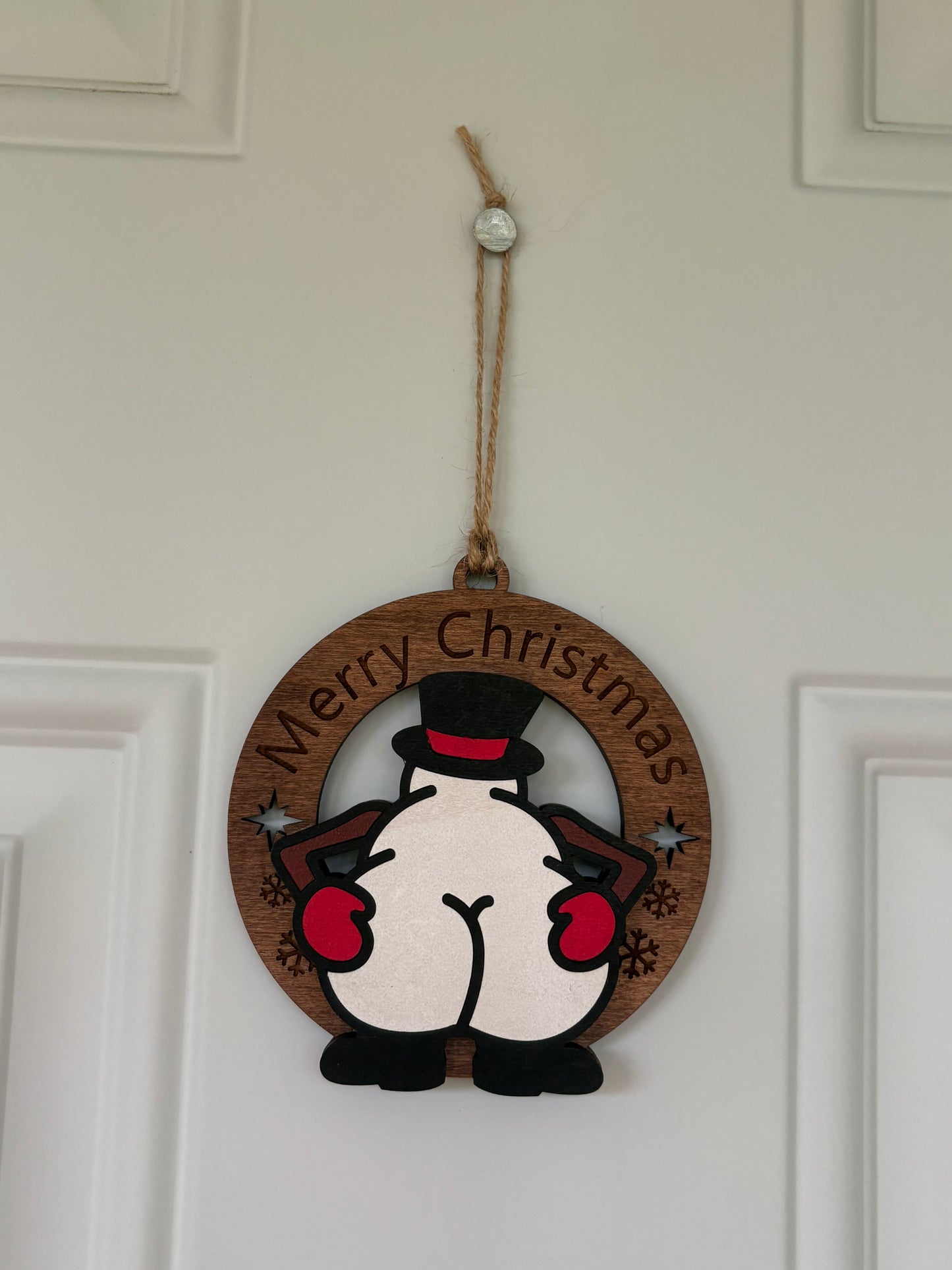 A round wooden Christmas hanging ornament featuring a cartoon penguin from behind with the words 'Merry Christmas' curved along the top edge, suspended by a jute string on a white paneled door.
