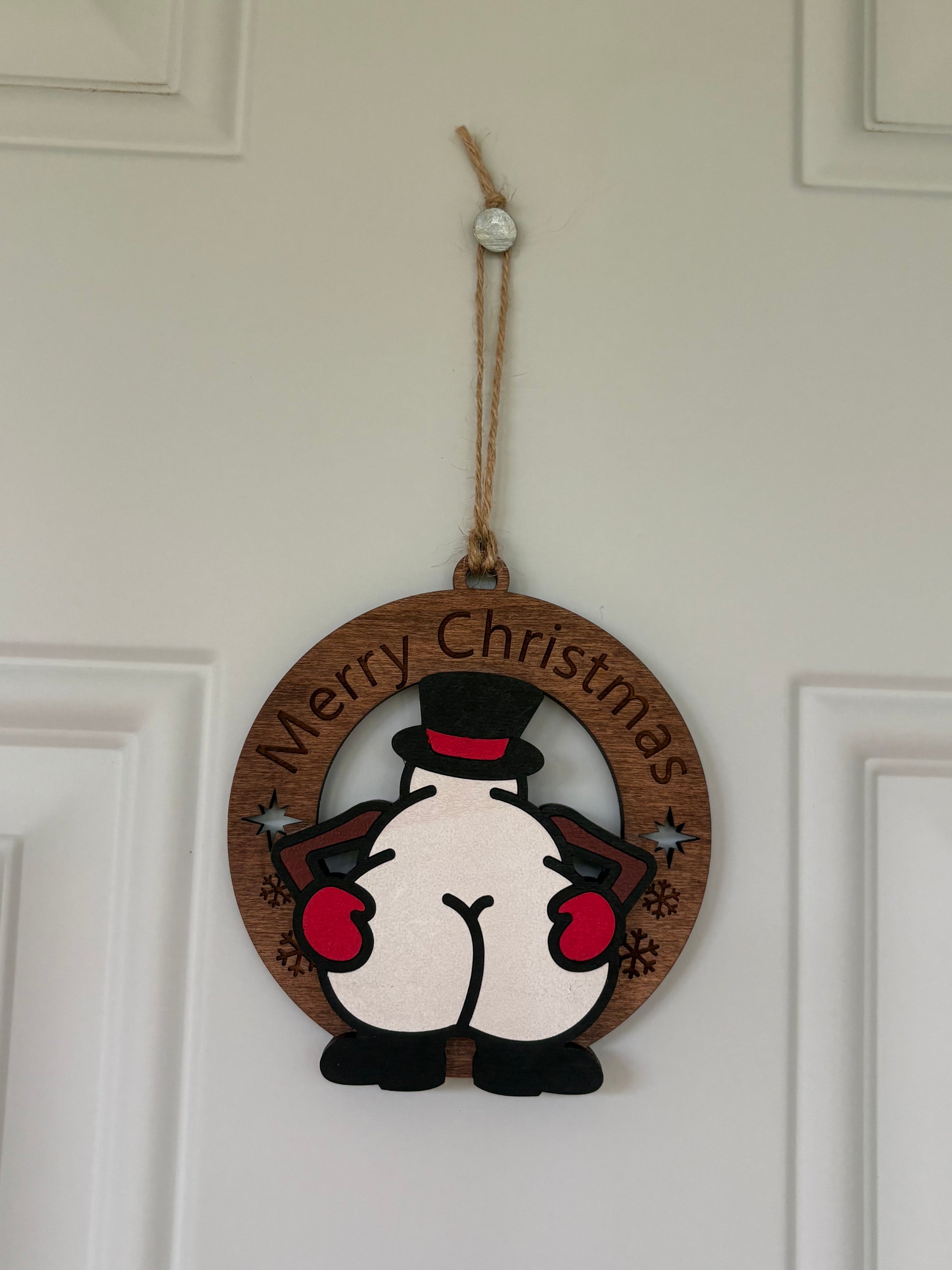 A round wooden Christmas hanging ornament featuring a cartoon penguin from behind with the words 'Merry Christmas' curved along the top edge, suspended by a jute string on a white paneled door.
