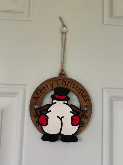 A round wooden Christmas hanging ornament featuring a cartoon penguin from behind with the words 'Merry Christmas' curved along the top edge, suspended by a jute string on a white paneled door.